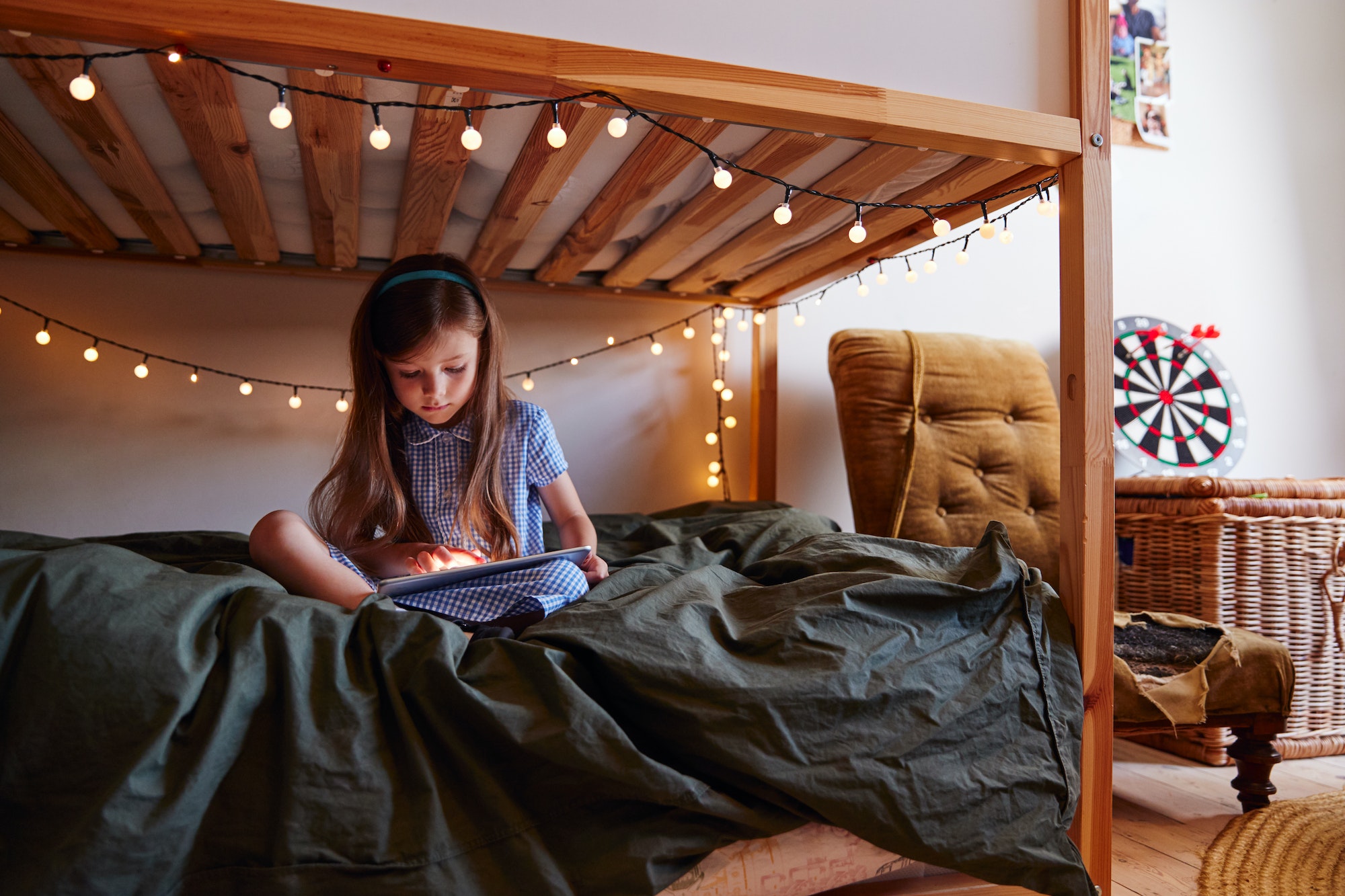 Young Girl Sitting On Bed At Home Decorated With Fairy Lights Using Digital Tablet